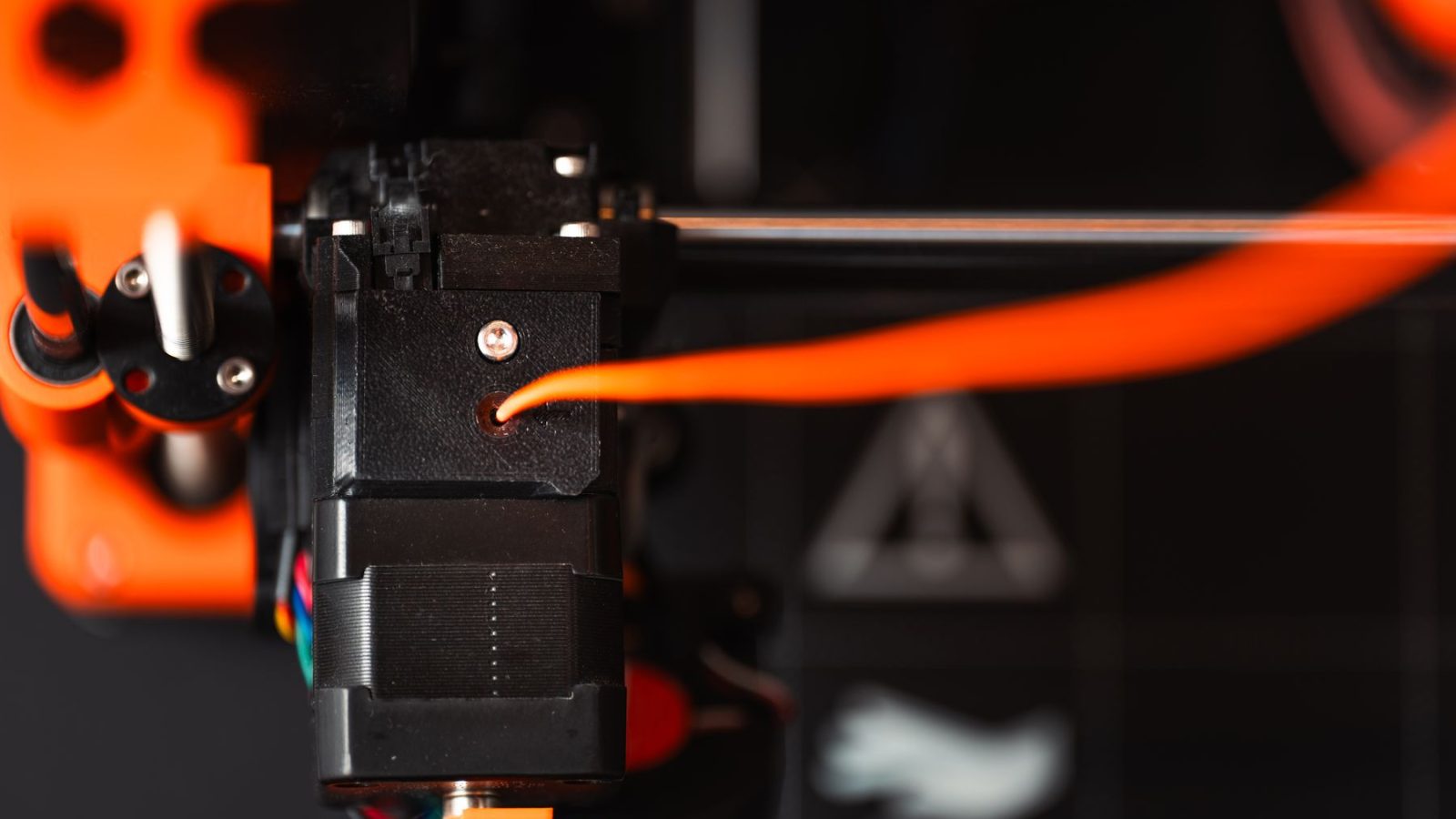 Podroben pogled na ekstruder 3D tiskalnika z oranžnim filamentom v industrijskem okolju