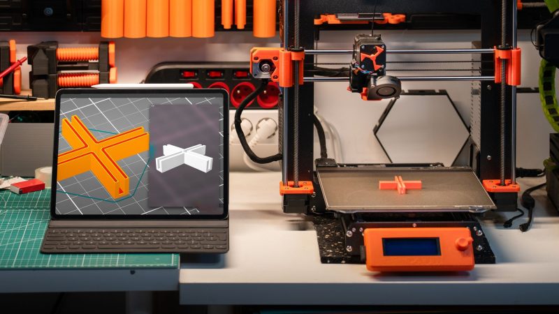 Arbeitsplatz mit 3D-Drucker und Computer, auf dem 3D-Modelle zu sehen sind, als Beispiel für moderne Technik im Einsatz
