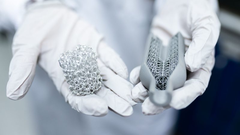 Laborumgebung mit behandschuhten Händen, die innovative 3D-gedruckte Metallstrukturen halten