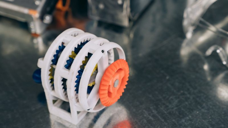 Nahaufnahme eines 3D-gedruckten Zahnradmechanismus in leuchtenden Farben auf einer spiegelnden Metalloberfläche
