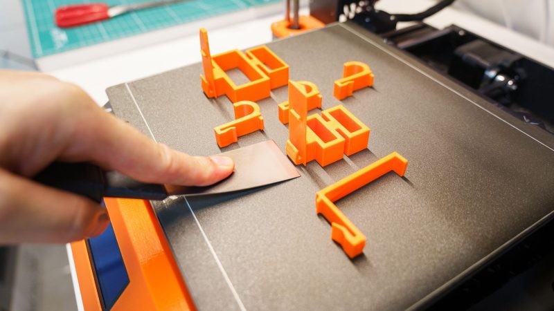 A hand carefully removes an orange 3D printed object from a printer bed using a spatula indoors